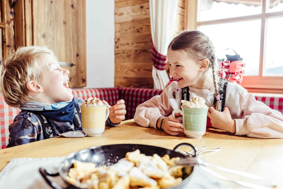 Zwei Kinder sitzen in der Skihütte, trinken heiße Schokolade mit Sahne und haben vor sich eine Pfanne voller Kaiserschmarrn stehen | © Alpbachtal Tourismus | shootandstyle 