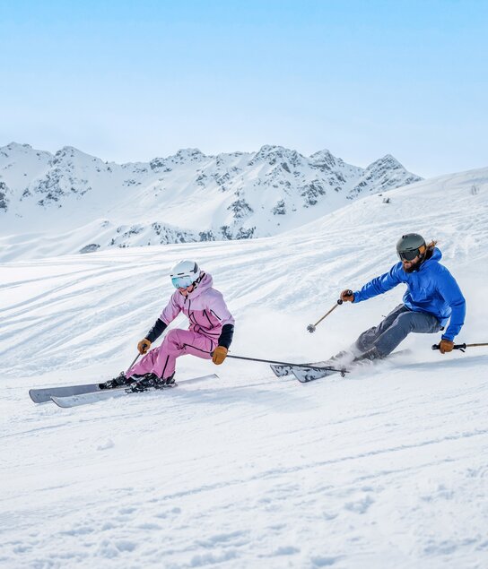 Ein Mann und eine Frau auf der Piste beim Skifahren mit Panorama im Hintergrund. | © Ski Juwel Alpbachtal Wildschönau | Christoph Oberschneider